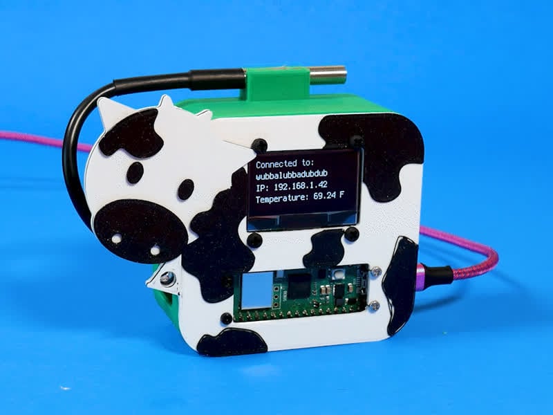 A Raspberry Pi Pico W HTTP server displaying information on an OLED display. All mounted in a black and white plastic case decorated like a cow. A DS18B20 temperature sensor is atop the case.