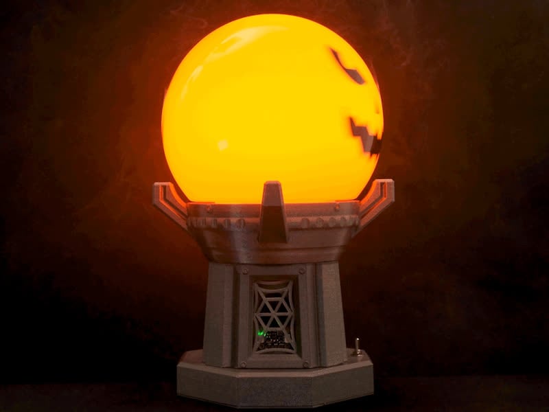 Glowing orange orb with a jack-o'-lantern face sits atop a gray 3D printed hexagonal base. A small green light is visible inside the base.